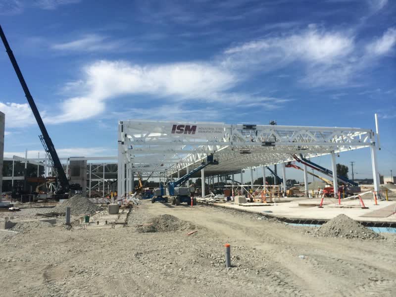 ISM banner hung on a white structural steel truss during erection of the Hamilton Transit Centre roof, with a crane and boom lifts working beneath a clear sky.