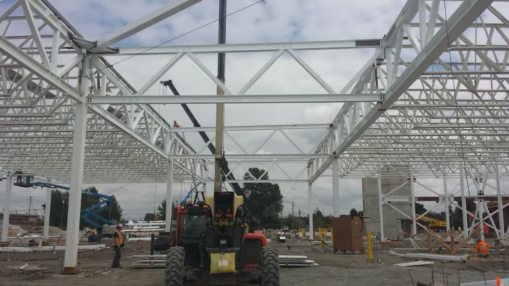 White steel roof trusses of the Hamilton Transit Centre overhead during erection, with a crane reaching through the frame.